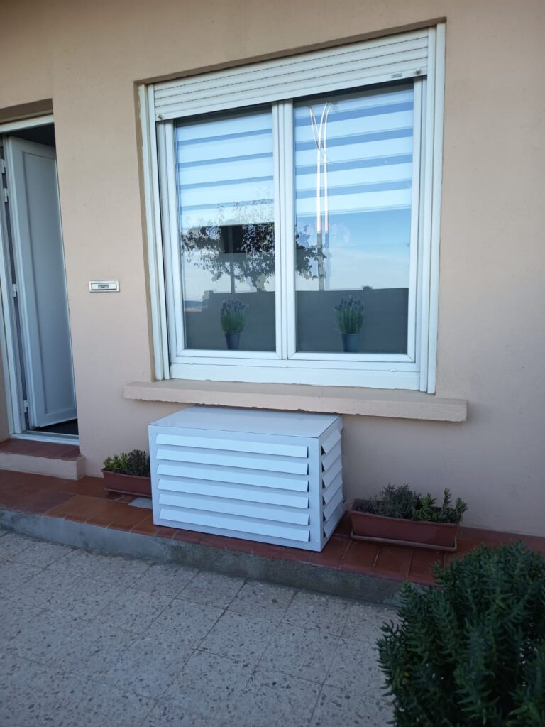 Cache-climatisation blanc à persiennes vu de côté, entre deux jardinières, à Canet-en-Roussillon